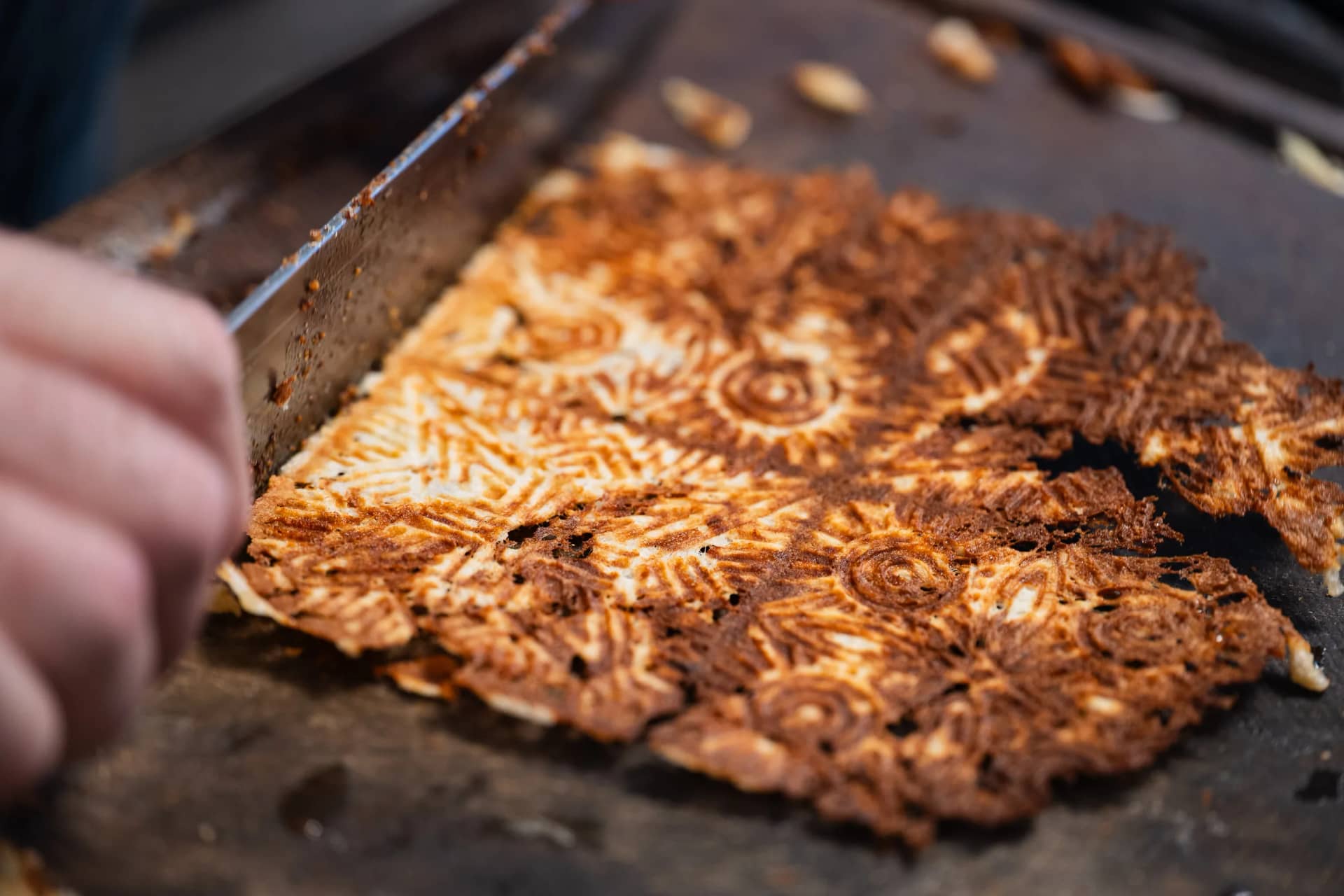 Une fine galette dorée, gravée des motifs du fer à bricelets, sur une planche sombre de la cuisine de la maison ; une lame de couteau en entame le bord