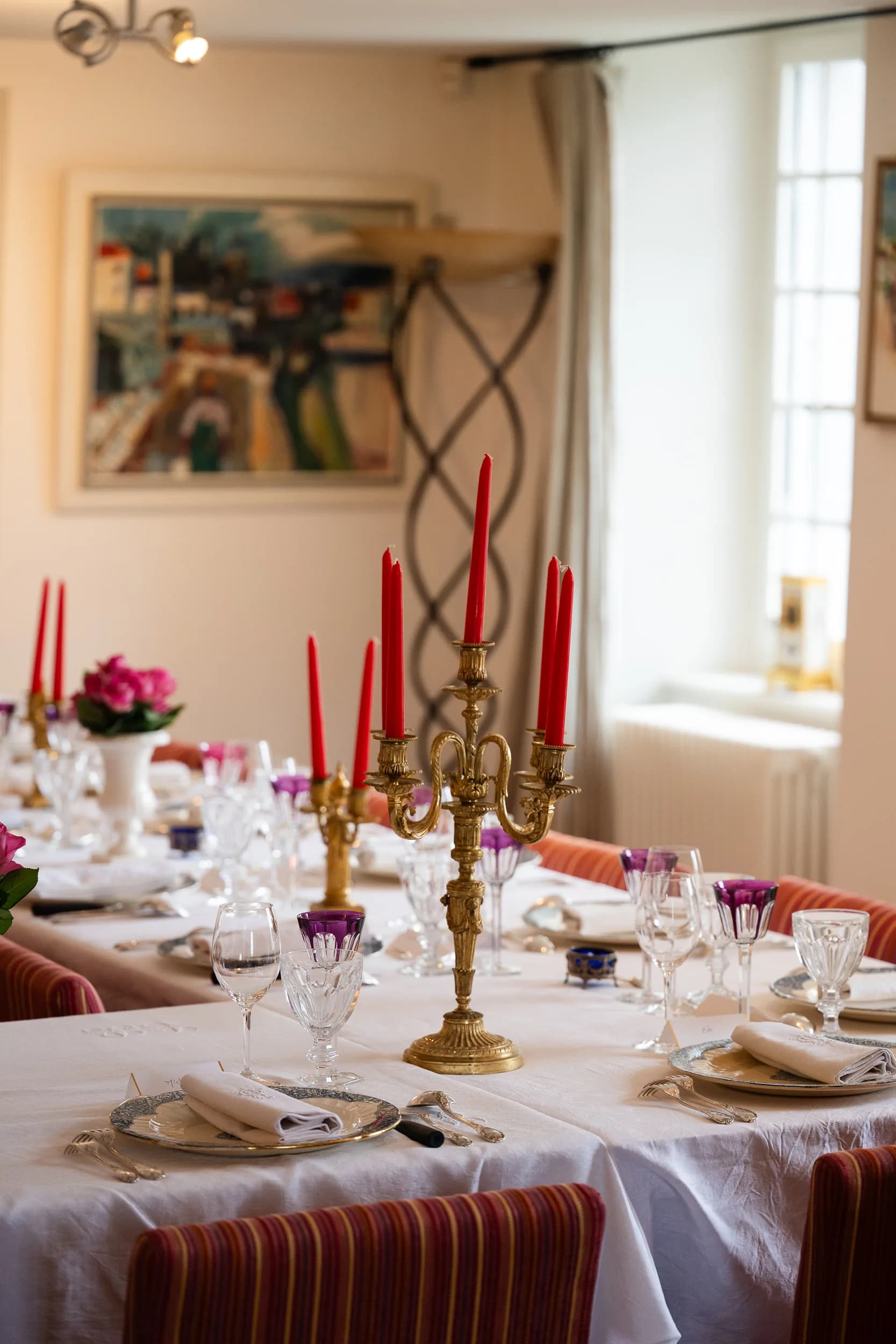 Un chandelier doré aux bougies rouges sur une table dressée, sous un tableau coloré et près d'une fenêtre de la salle à manger
