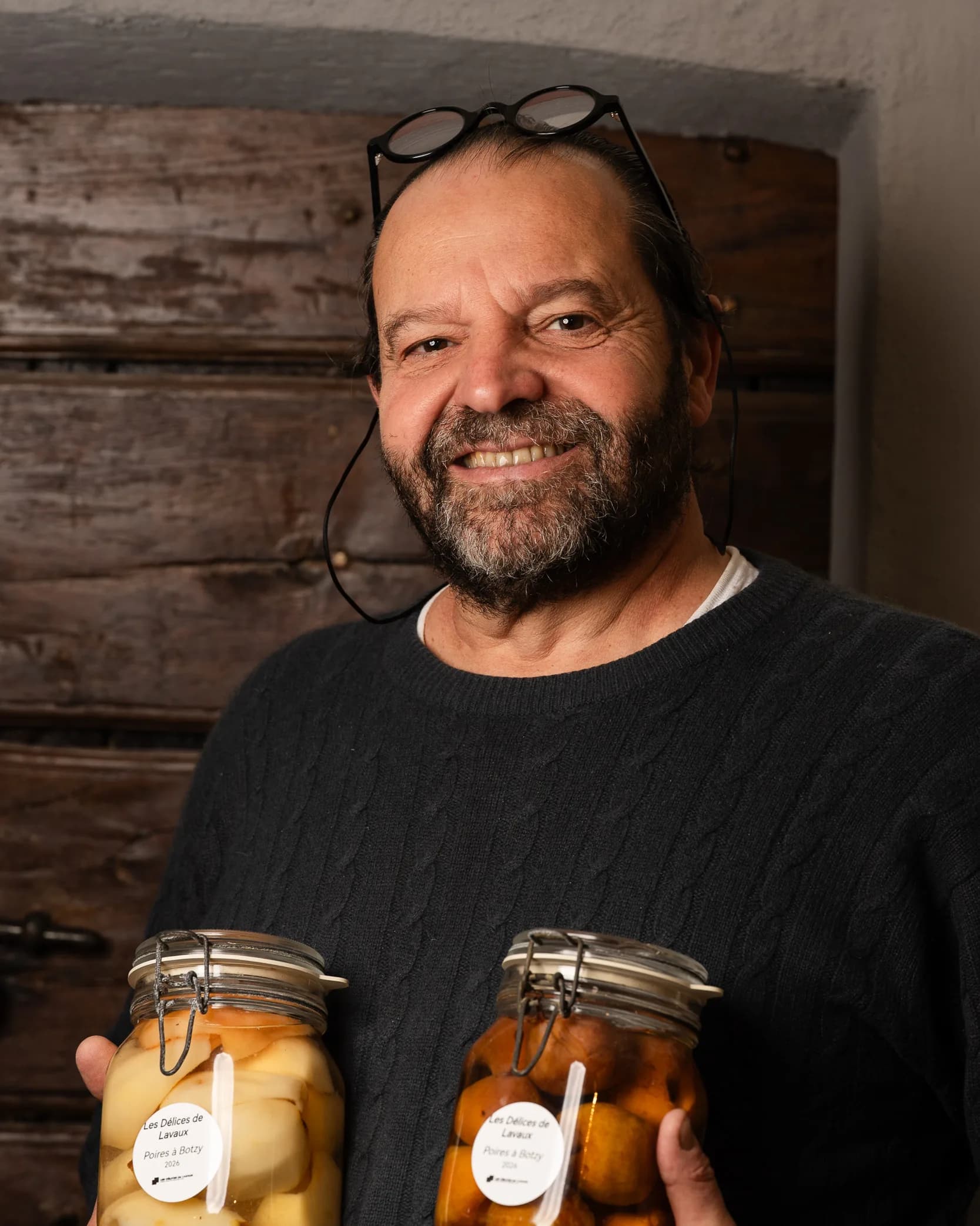 Andres Oppenheim sourit, ses lunettes relevées sur la tête, deux grands bocaux de poires au sirop dans les mains, devant une vieille porte en bois