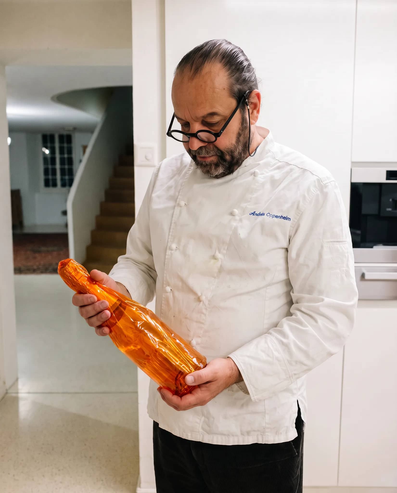 Andres Oppenheim en veste de chef blanche tient une bouteille de champagne dans son emballage orange, dans le hall clair de la maison, l'escalier derrière lui