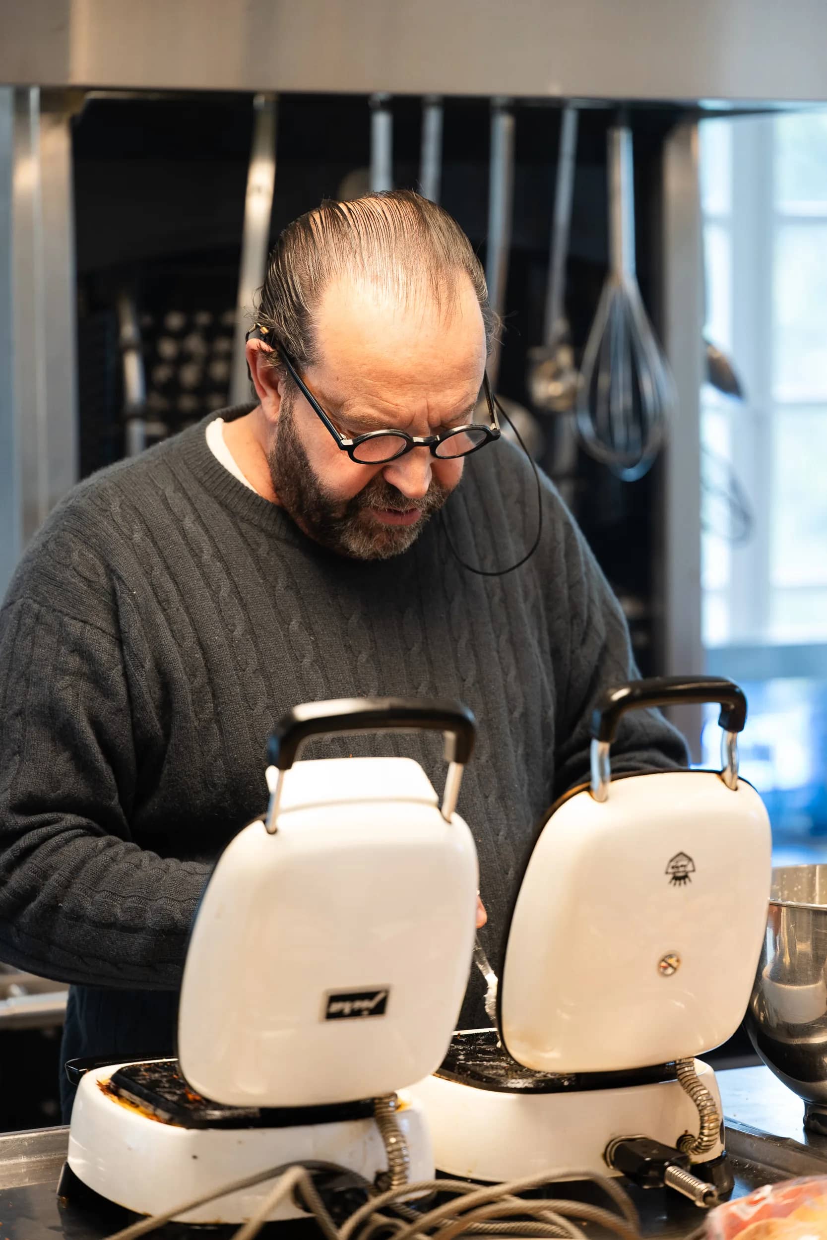 Andres Oppenheim, en pull sombre, au-dessus de deux fers à bricelets ouverts, devant les ustensiles suspendus de sa cuisine