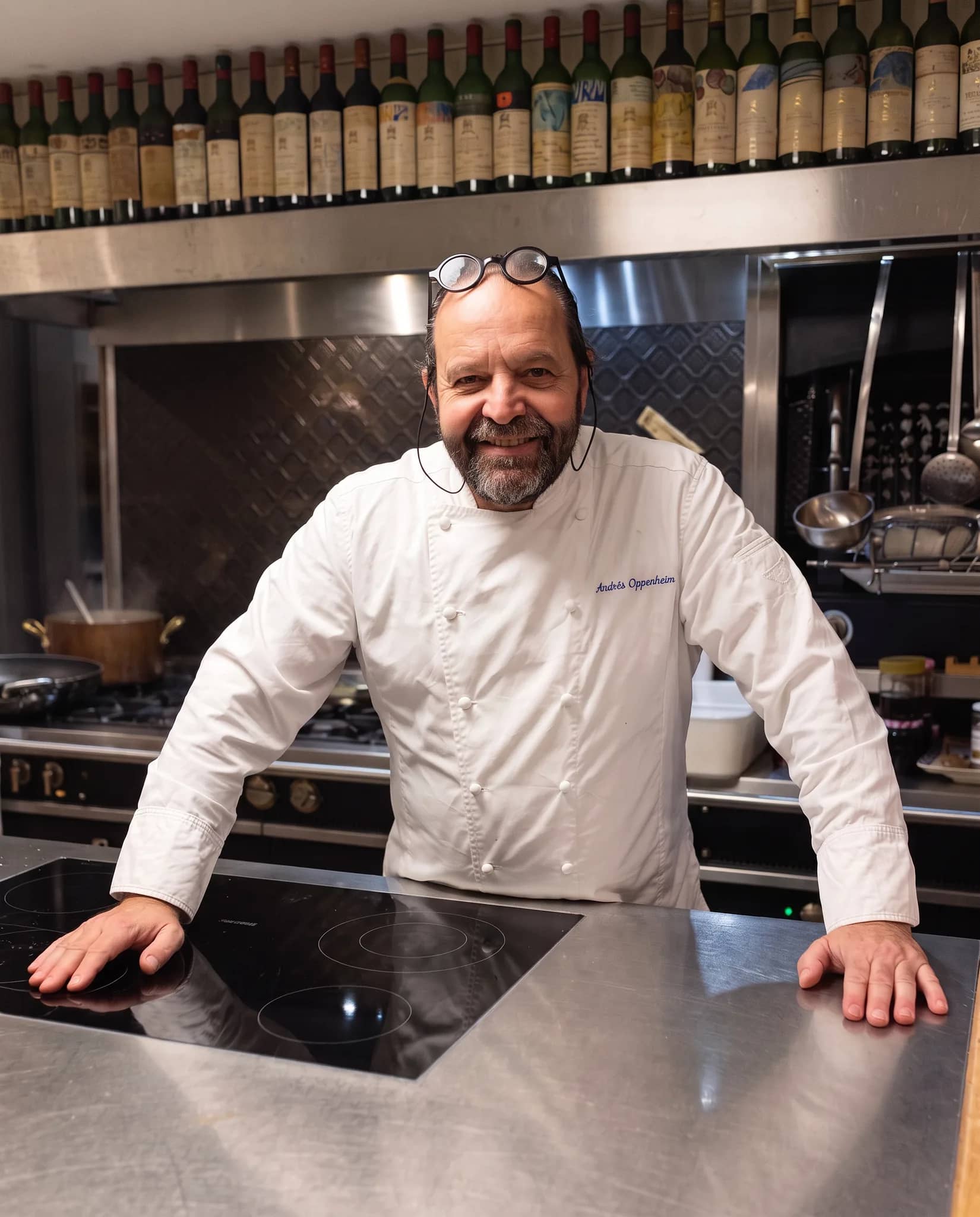 Andres Oppenheim en veste de chef blanche, les deux mains posées sur l'îlot en inox de sa cuisine, sourit, ses lunettes relevées sur la tête, sous une longue rangée de vieilles bouteilles