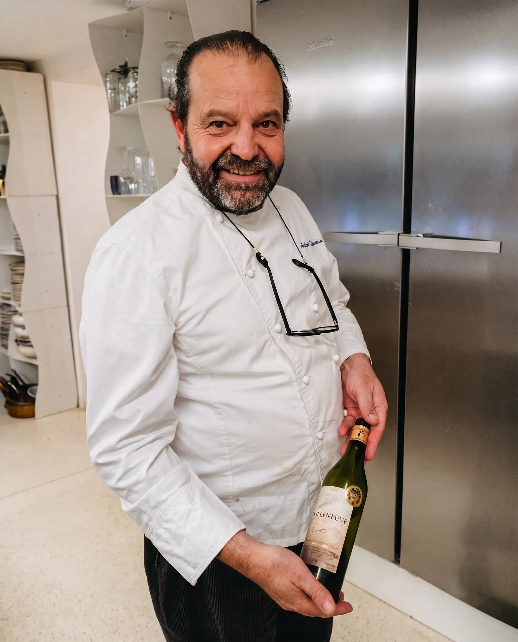Andres Oppenheim en veste de chef blanche, ses lunettes pendues à leur cordon, tient une bouteille de vin blanc et sourit, près du grand réfrigérateur en inox de sa cuisine