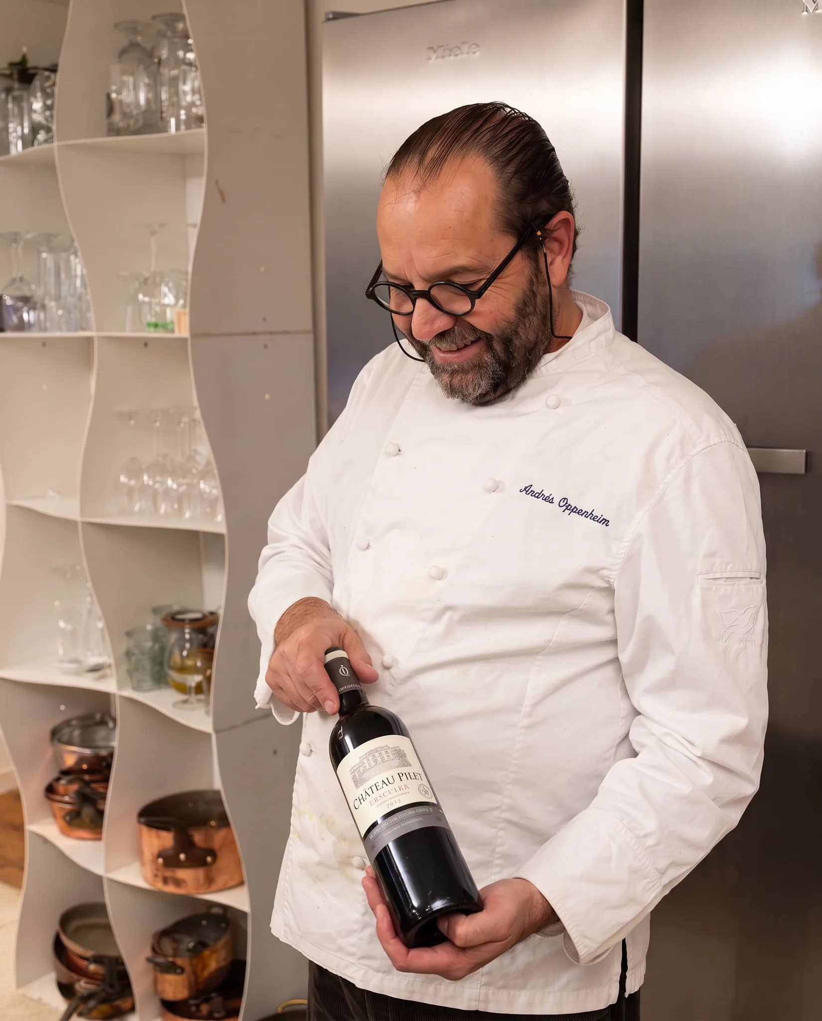 Andres Oppenheim en veste de chef blanche présente une bouteille de vin rouge et en regarde l'étiquette en souriant, dans sa cuisine aux étagères blanches