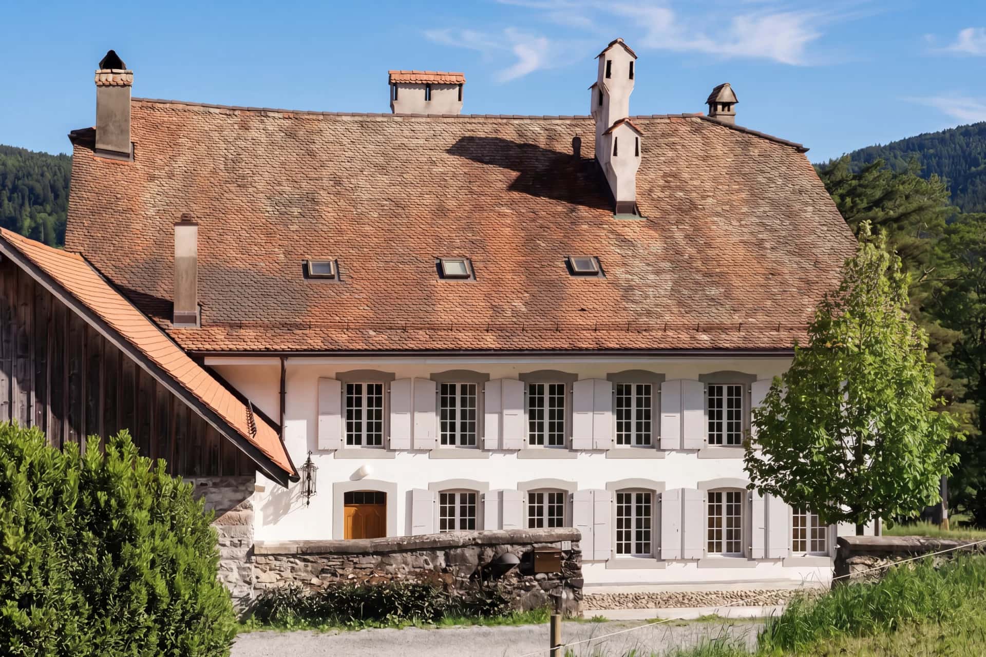 La façade côté cour de la maison de maître de Puidoux : volets gris, grand toit de tuiles anciennes et cheminées, la grange à gauche, le mur de pierre et le chemin de gravier