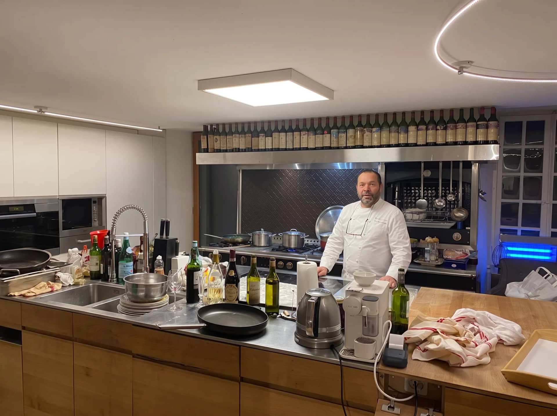 La cuisine de la maison un soir : l'îlot et son plan de travail en bois chargé de bouteilles et de casseroles, et Andres Oppenheim en veste blanche au fourneau, sous l'étagère des vieilles bouteilles