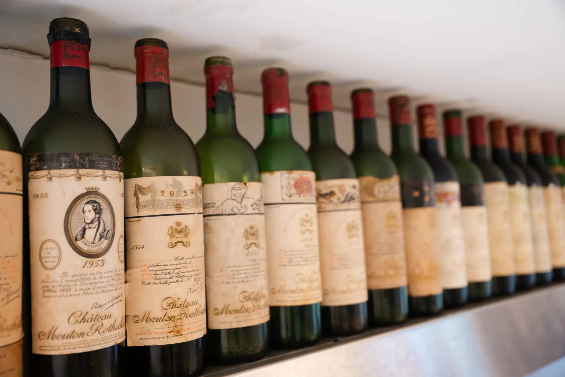 Une longue rangée de grands bordeaux anciens aux étiquettes d'artistes, alignés sur une étagère de la cuisine
