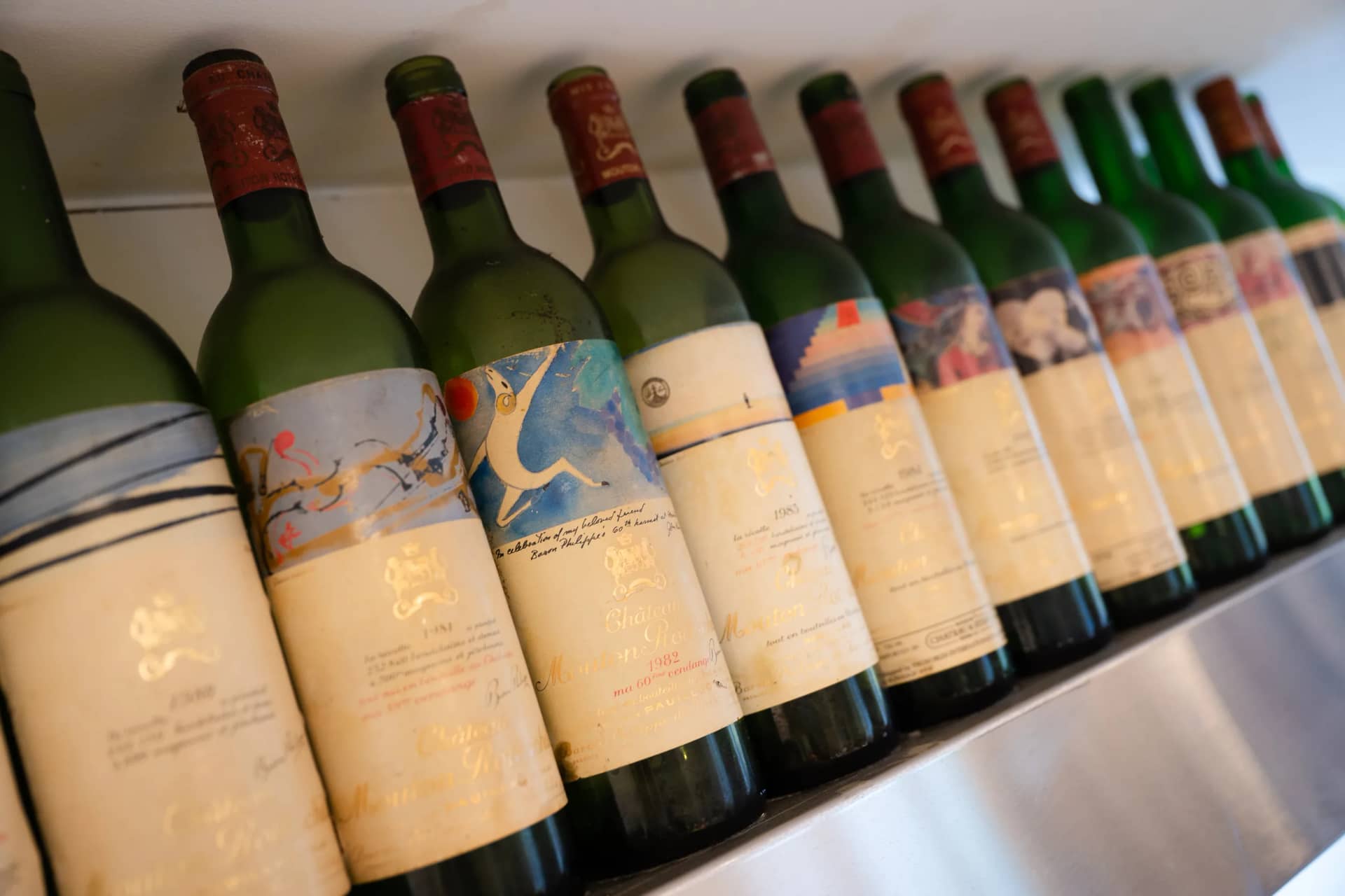 Des grands bordeaux anciens en rang sur une étagère de la cuisine, leurs étiquettes peintes par des artistes au premier plan
