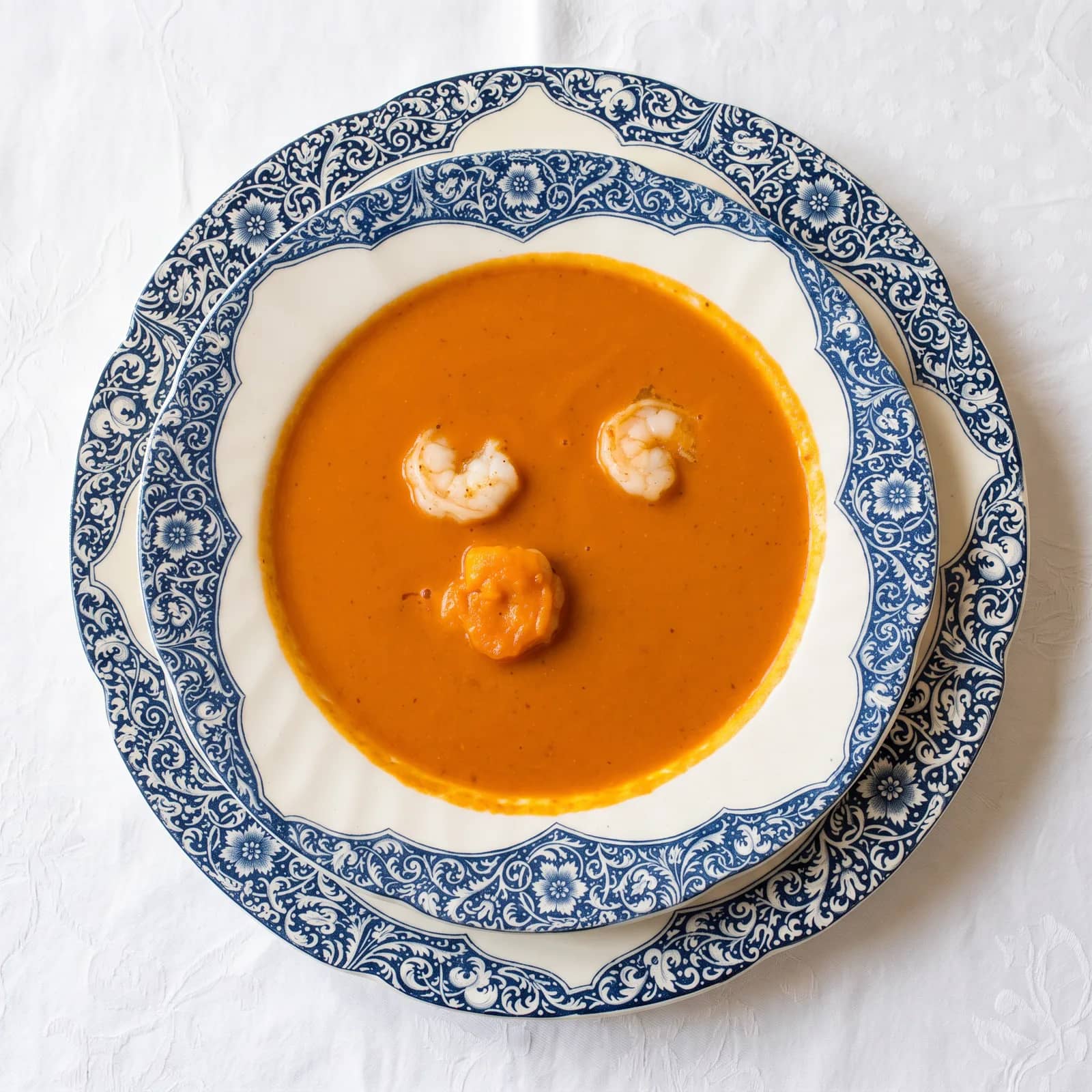 Vue de dessus, une bisque de crustacés orangée et trois crevettes dans une assiette creuse en porcelaine à frise bleue, sur la nappe damassée blanche