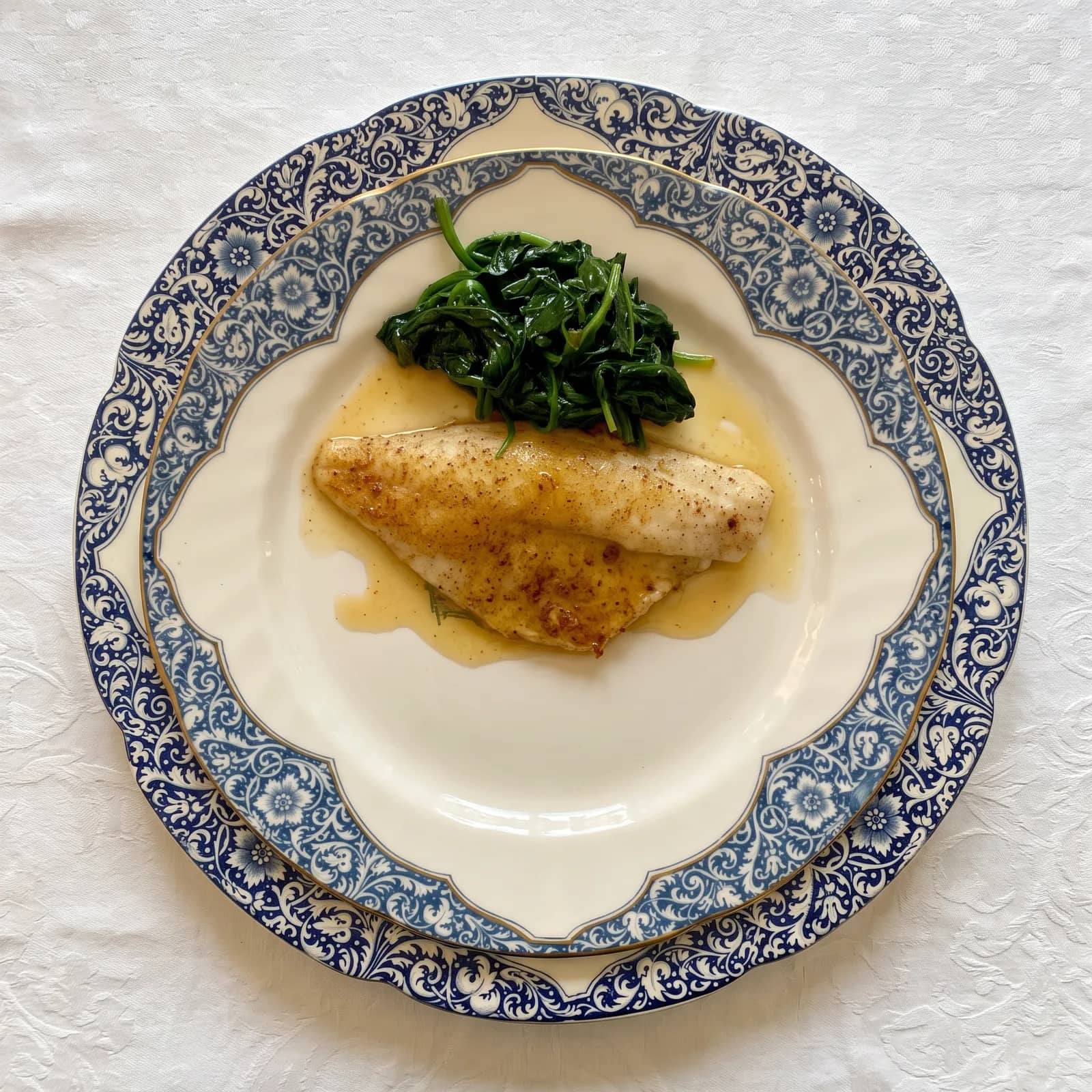 Vue de dessus, un filet de poisson doré sur des épinards, nappé de beurre, dans une assiette en porcelaine à frise bleue, sur la nappe damassée blanche