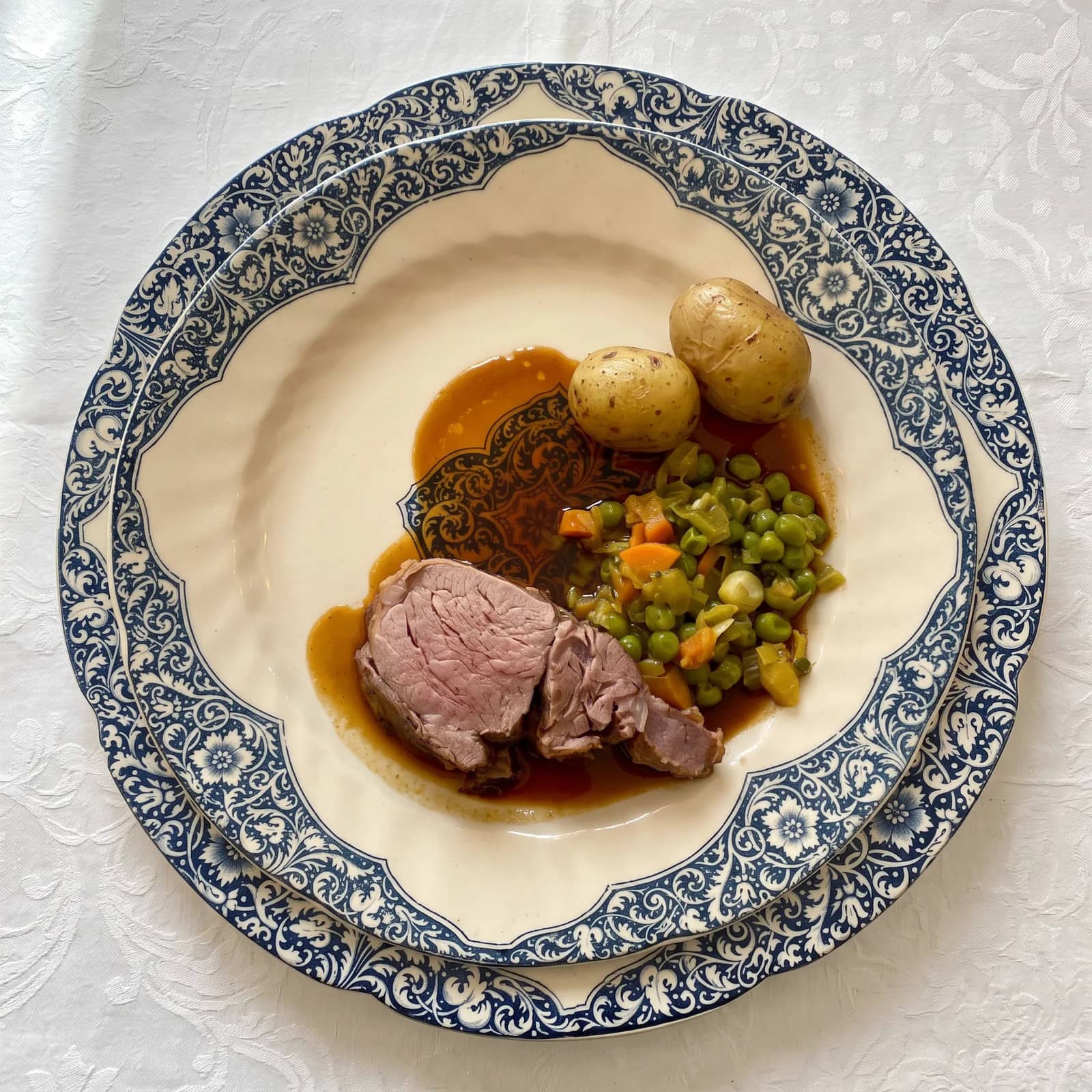 Vue de dessus, des tranches de viande rosée et leur jus, des pommes de terre nouvelles et des petits pois aux carottes, dans une assiette en porcelaine à frise bleue