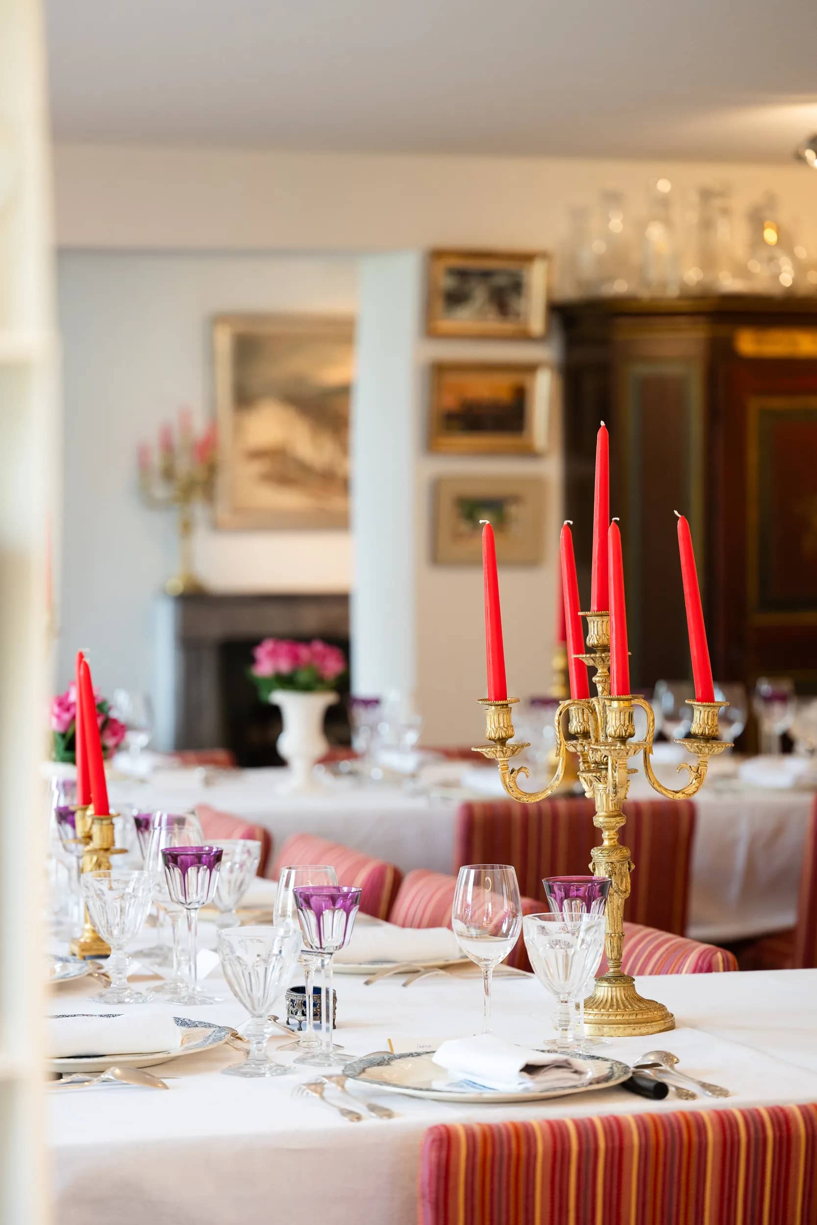 Le grand chandelier doré aux cinq bougies rouges et des verres en cristal améthyste au premier plan ; au fond, la salle à manger, ses tableaux, un bouquet de roses et une armoire peinte