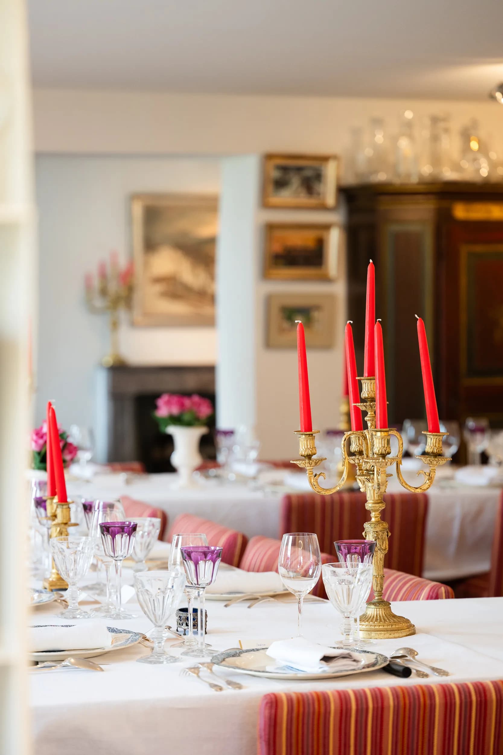 Le grand chandelier doré aux cinq bougies rouges et des verres en cristal améthyste au premier plan ; au fond, la salle à manger, ses tableaux, un bouquet de roses et une armoire peinte