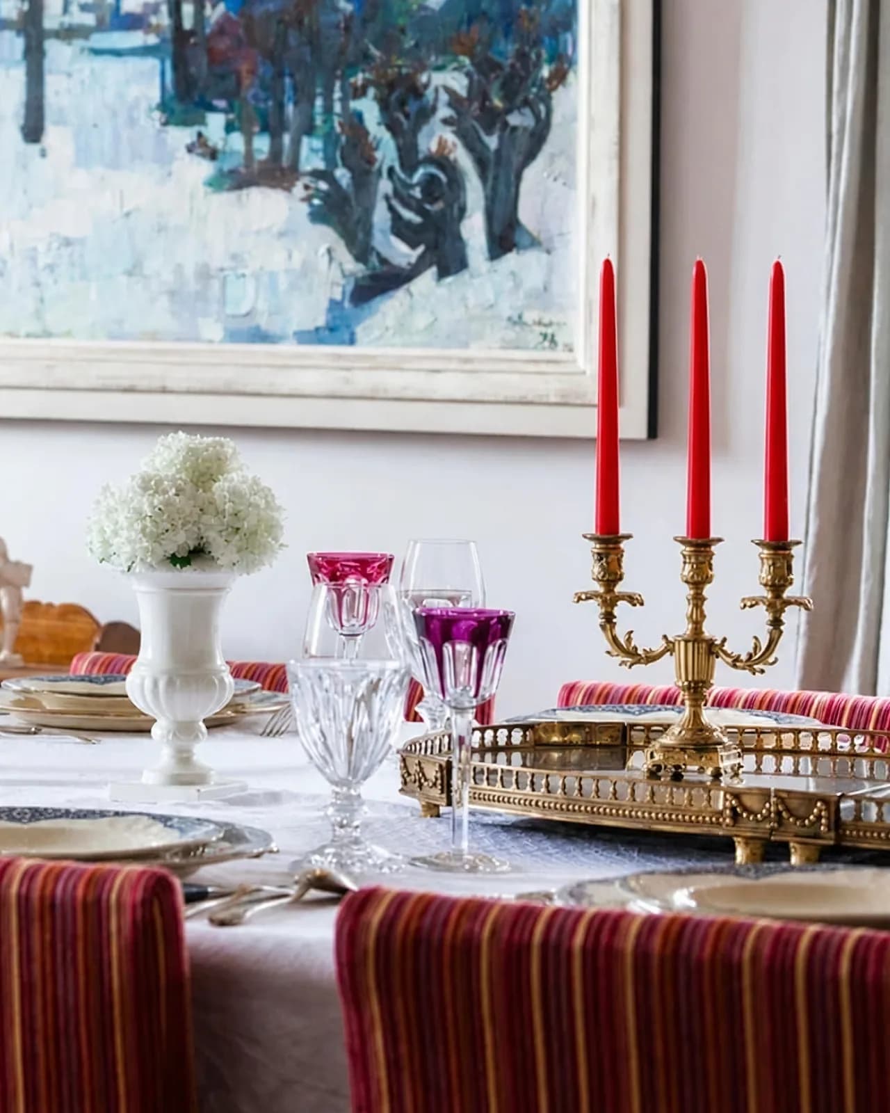 La même table à la lumière du jour : chandelier doré à trois bougies rouges, verres en cristal coloré et bouquet blanc devant un grand tableau