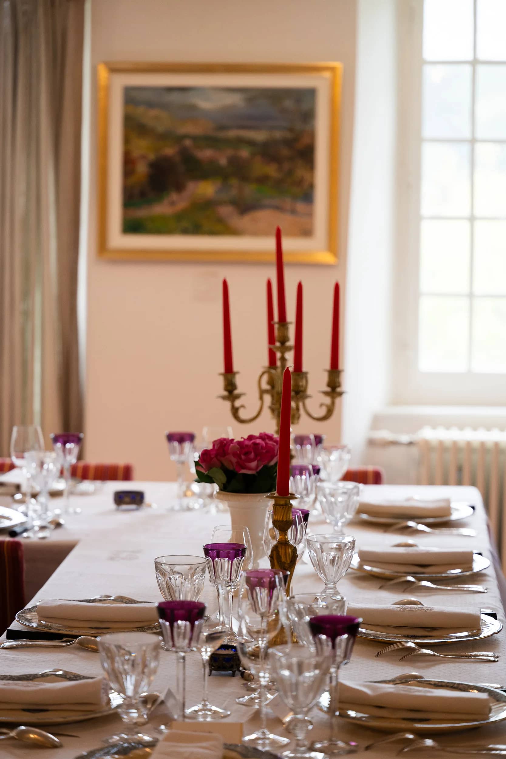 Un chandelier doré aux bougies rouges et un bouquet de roses sur la table de la salle à manger, sous un paysage encadré