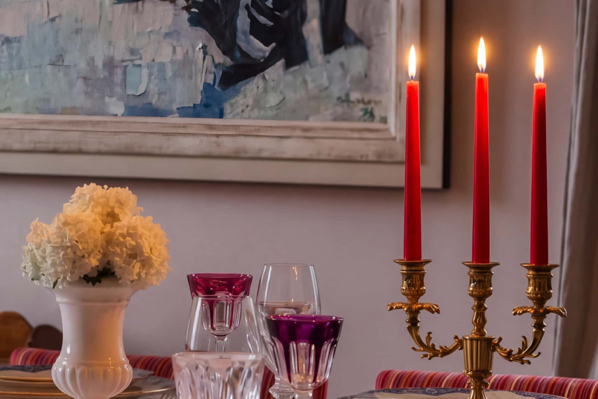 Un chandelier doré aux trois bougies rouges allumées et des verres en cristal taillé, rose et améthyste, sur la table de la salle à manger, sous un tableau