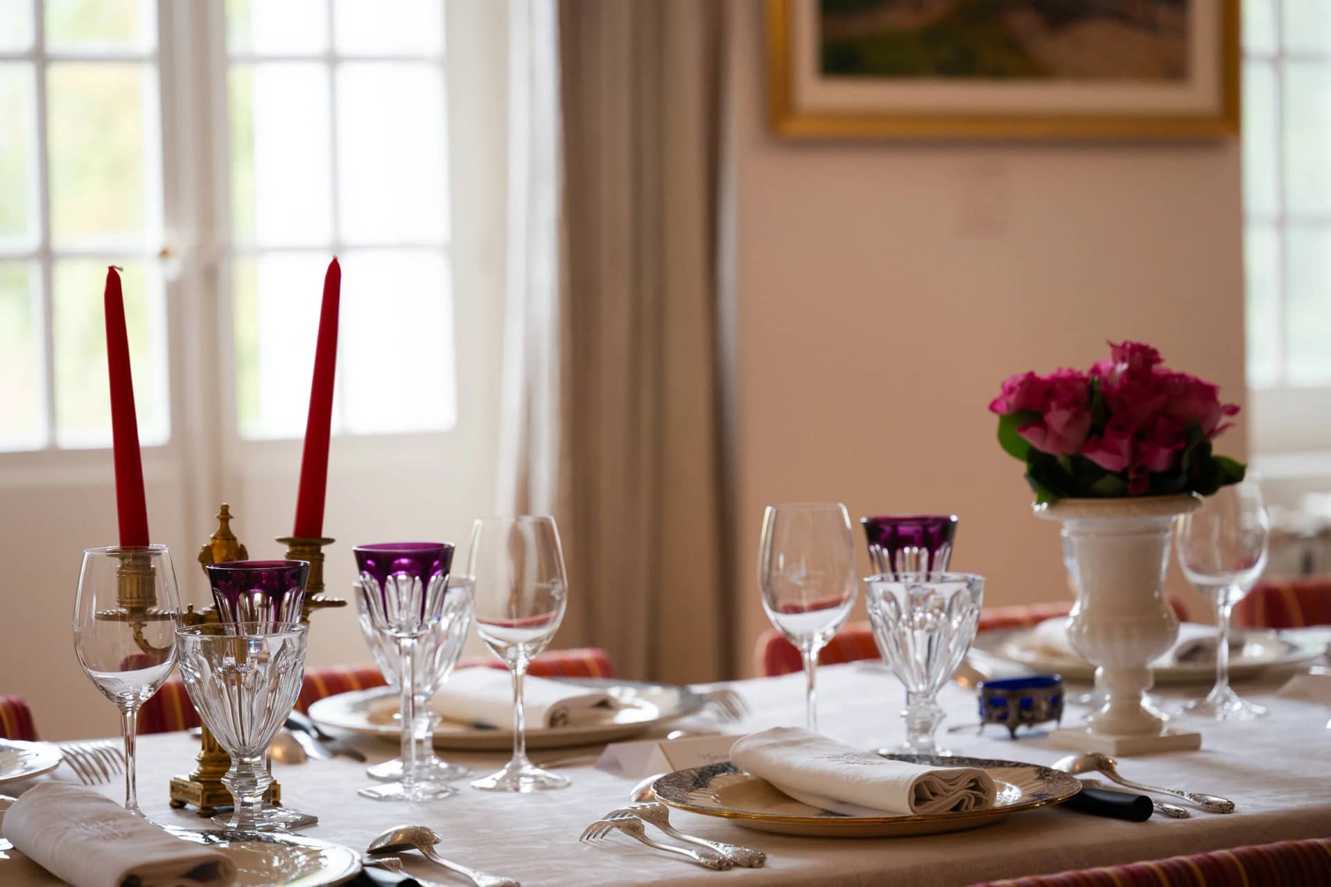 Une table dressée devant les portes-fenêtres de la salle à manger : bougies rouges, verres en cristal taillé améthyste et transparents, roses dans une vasque blanche, serviettes brodées