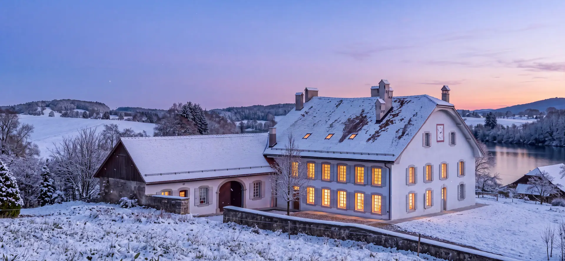 La maison de maître de Puidoux un soir de décembre, sous la neige, ses fenêtres éclairées, le Lac de Bret à l'arrière-plan